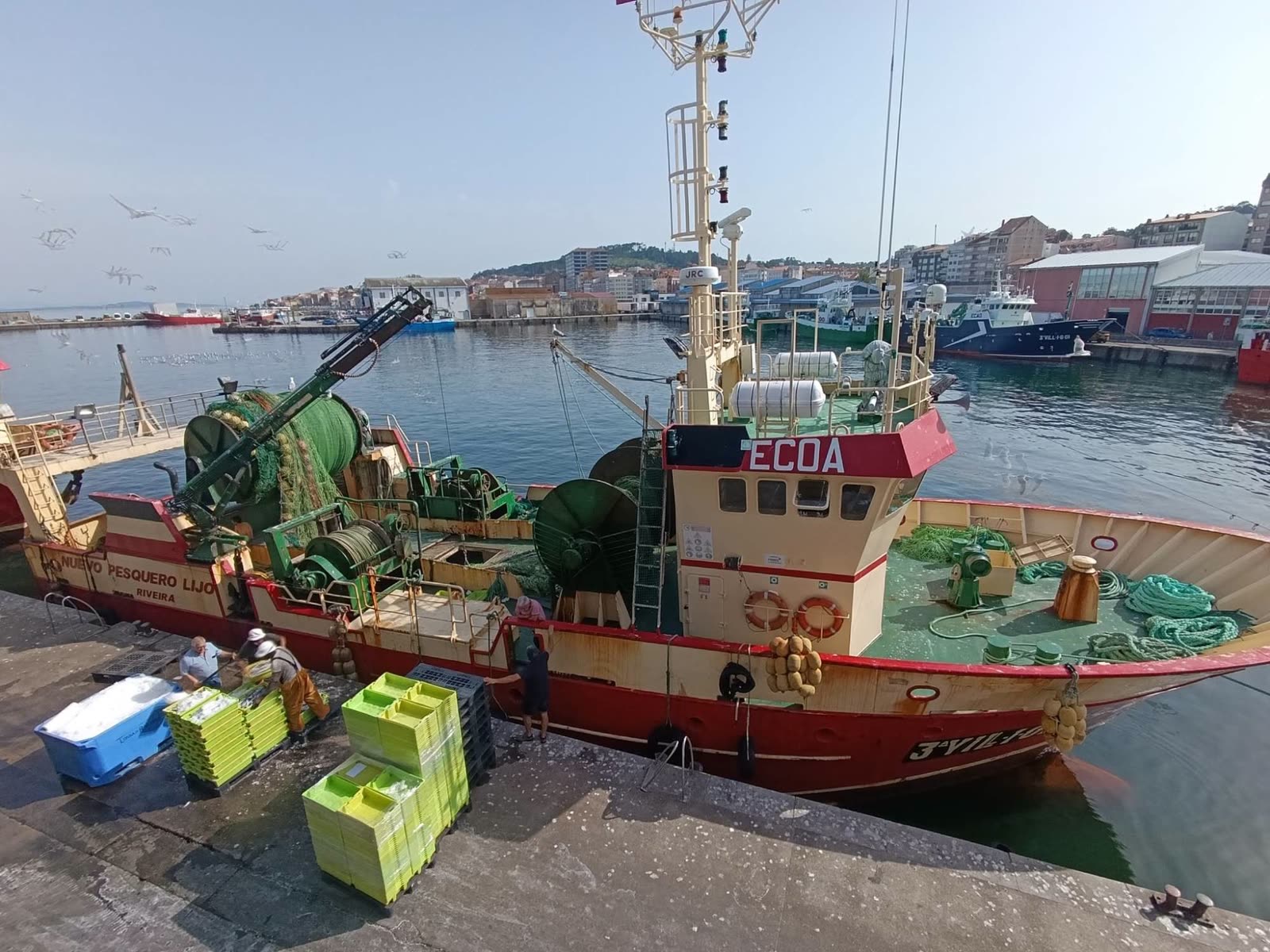 Mariñeiros descargando caixas de peixe dun arrastreiro no peirao de Ribeira, coa vila ao fondo