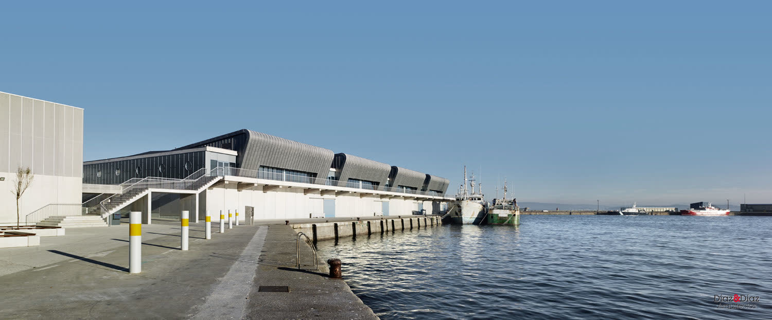Panorámica arquitectónica da lonxa de Ribeira sobre a auga, con barcos amarrados no peirao